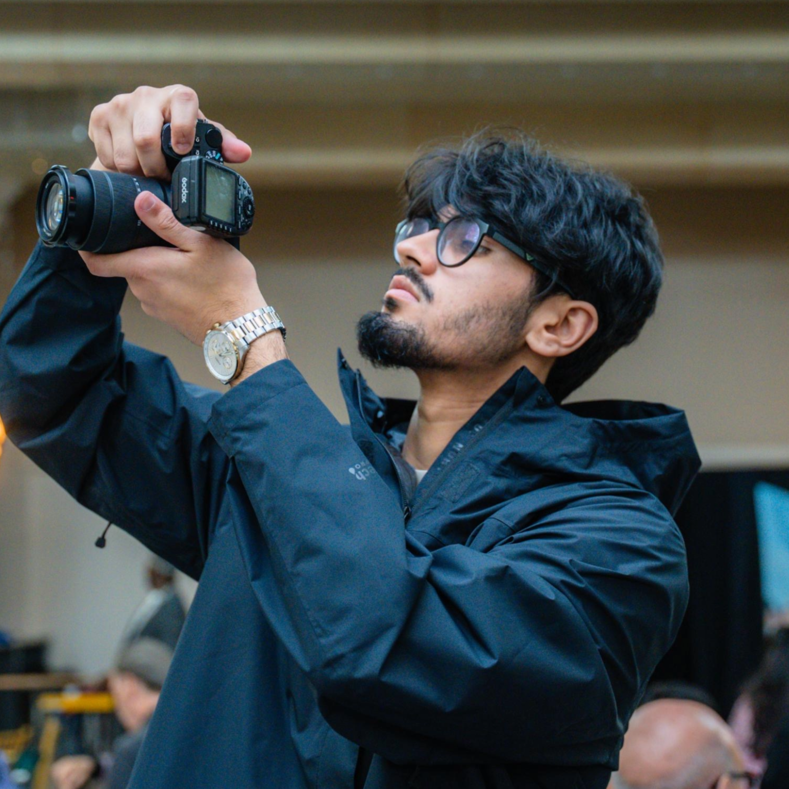 A man with black curly hair and glasses wearing a dark blue jacket is holding a camera up to take a photo, with a serious expression on his face.