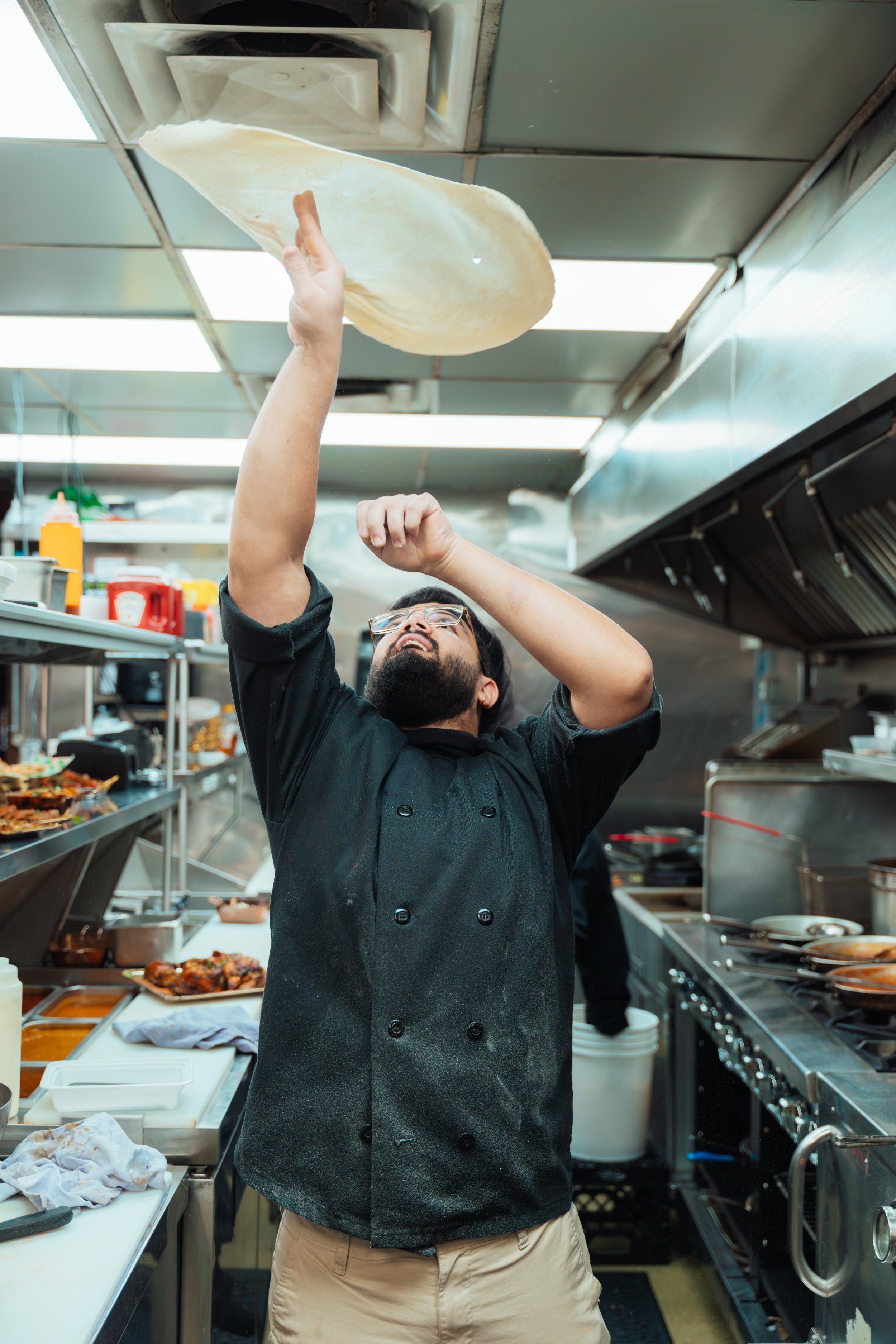 A chef tossing pizza dough in the air inside a commercial kitchen, with shelves of food and kitchen appliances visible in the background.