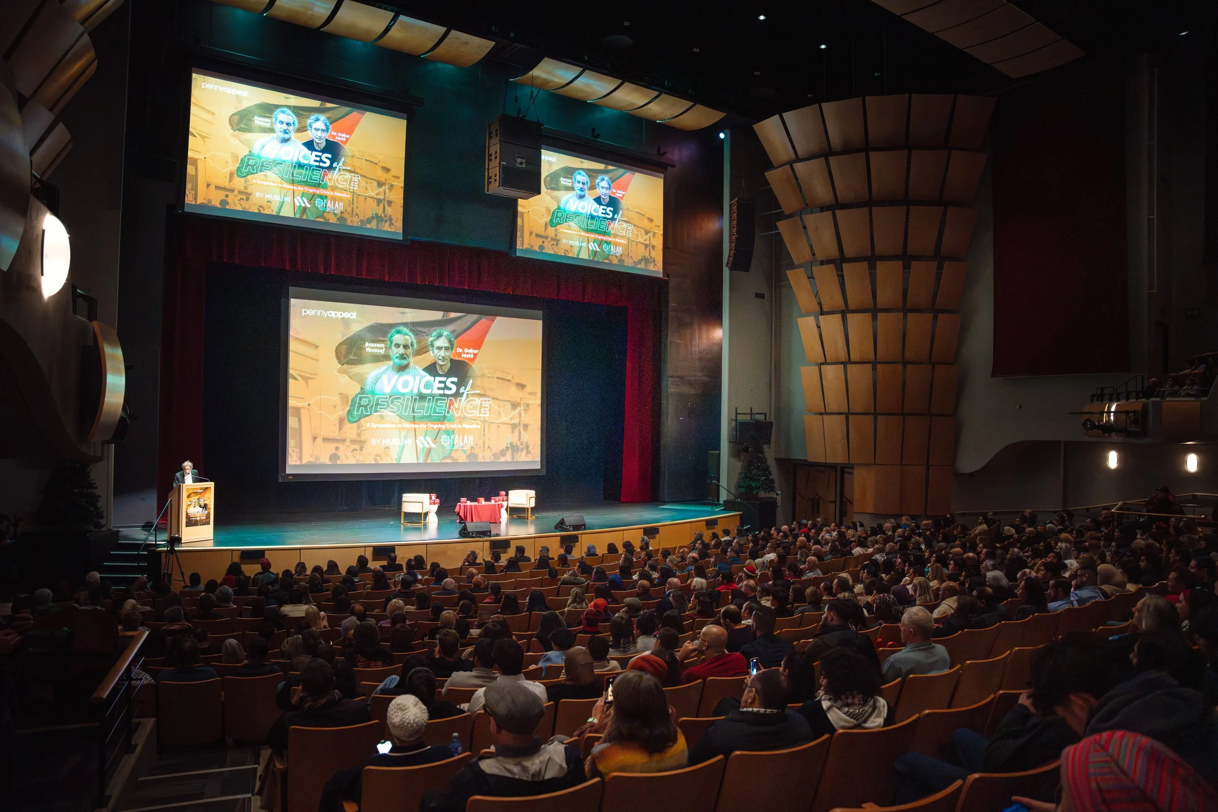 A large auditorium filled with people watching a stage with two large screens showing a presentation titled "Voices of Resilience." The stage has a podium and a small table with chairs, and the auditorium has wooden acoustic panels on the walls.