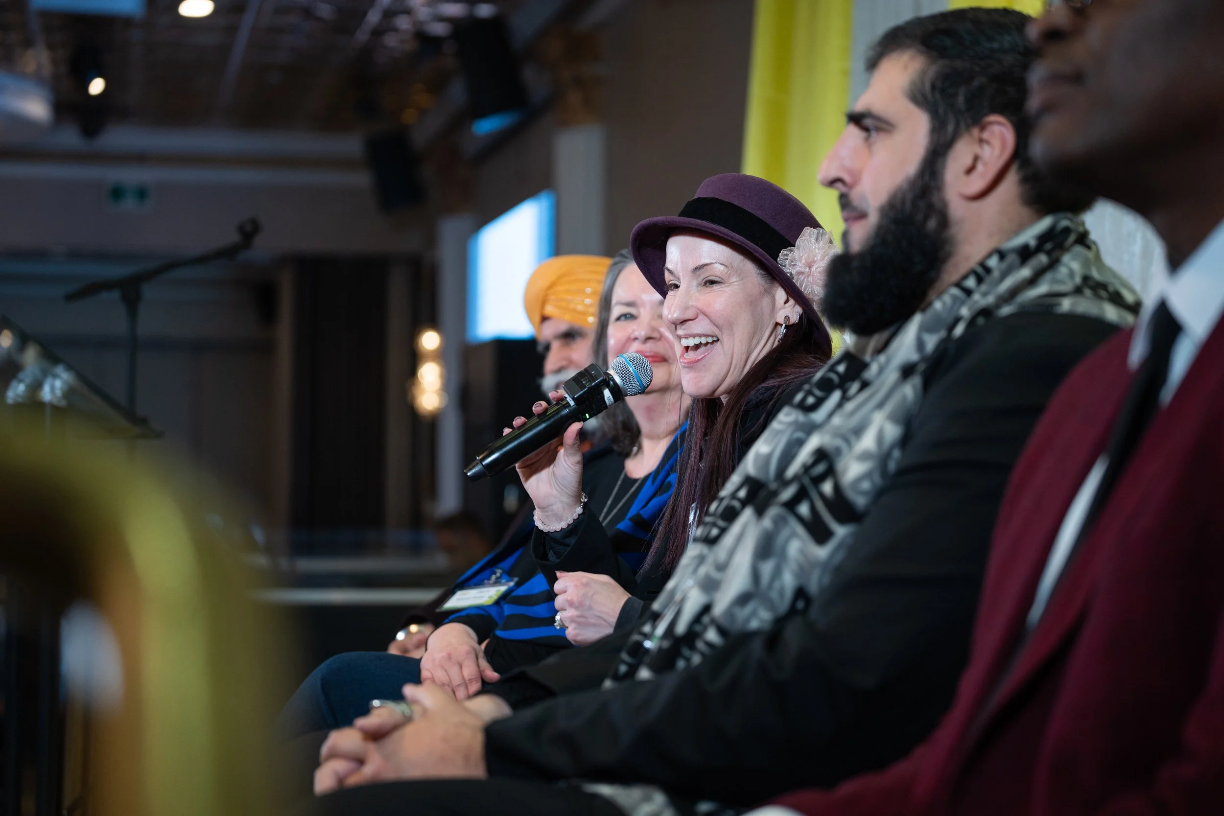 A group of diverse people sitting in a row at a conference or panel discussion. A woman in the middle is smiling and holding a microphone.