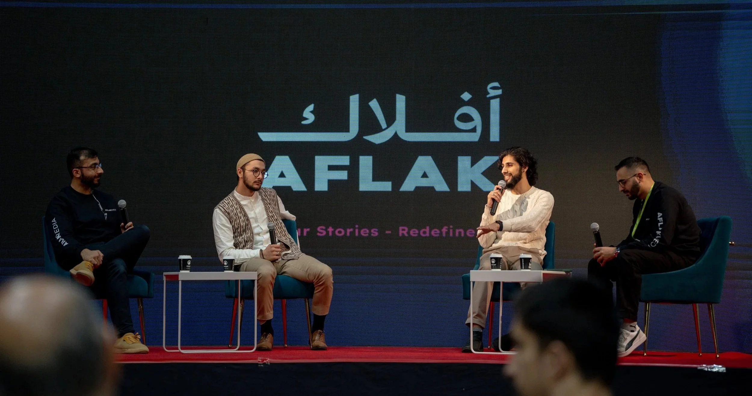 Four men seated on stage in front of a large screen with logo and the word 'AFLAK' during a panel discussion or presentation.