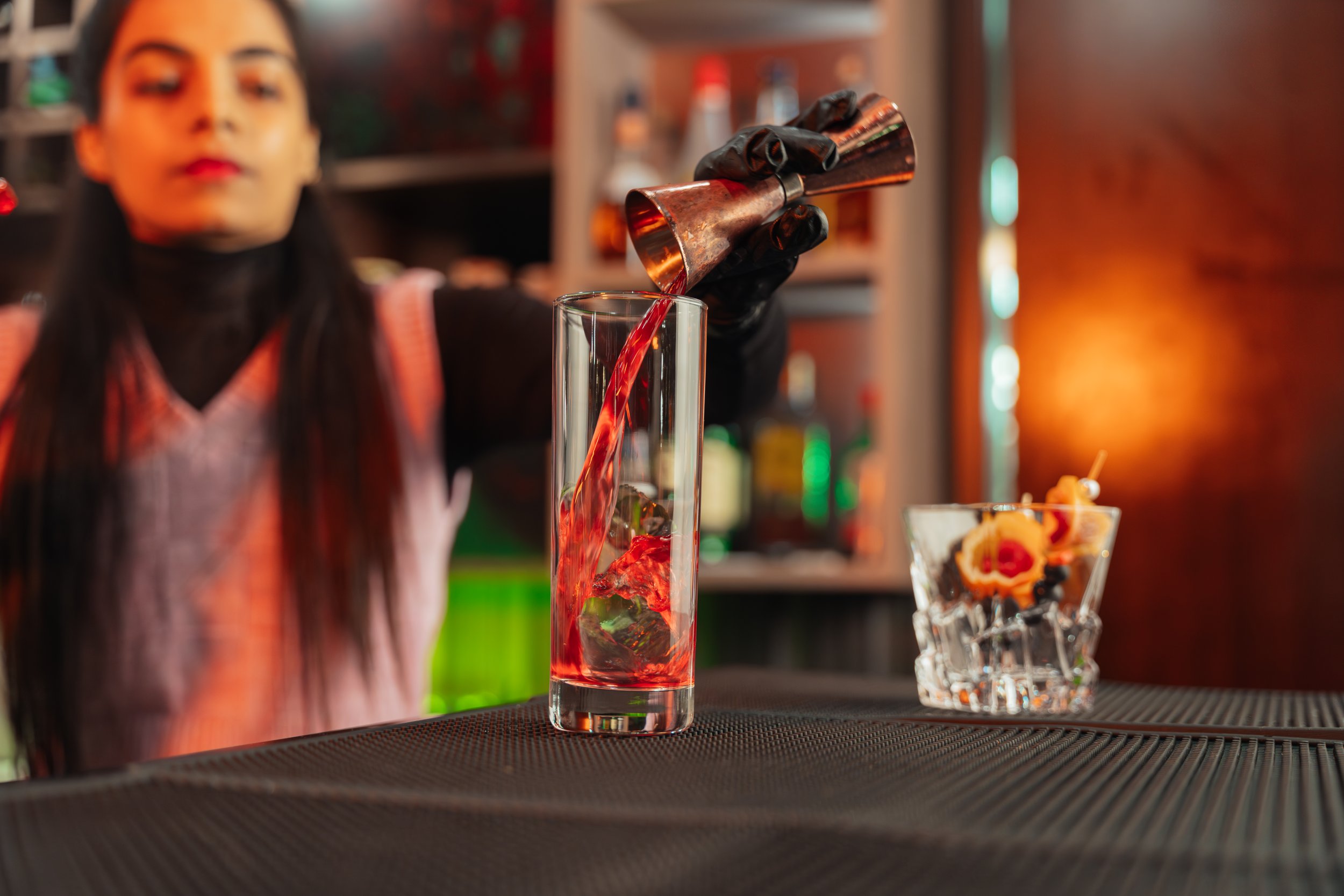 A woman with dark hair and red lipstick pours red liquid from a copper cocktail shaker into a tall glass with ice. In the background, a decorative glass with fruit garnishes is visible. The scene is in a bar setting with warm, ambient lighting.