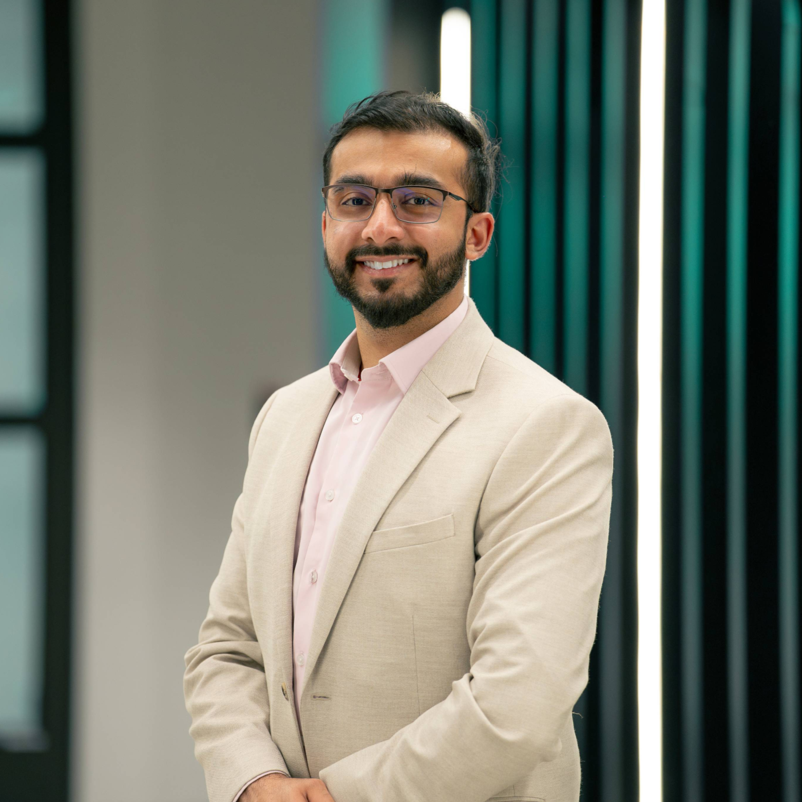 A young man with glasses, beard, and mustache, dressed in a light beige blazer and pink shirt, smiling and standing with arms crossed in front of a modern, neon-lit background.