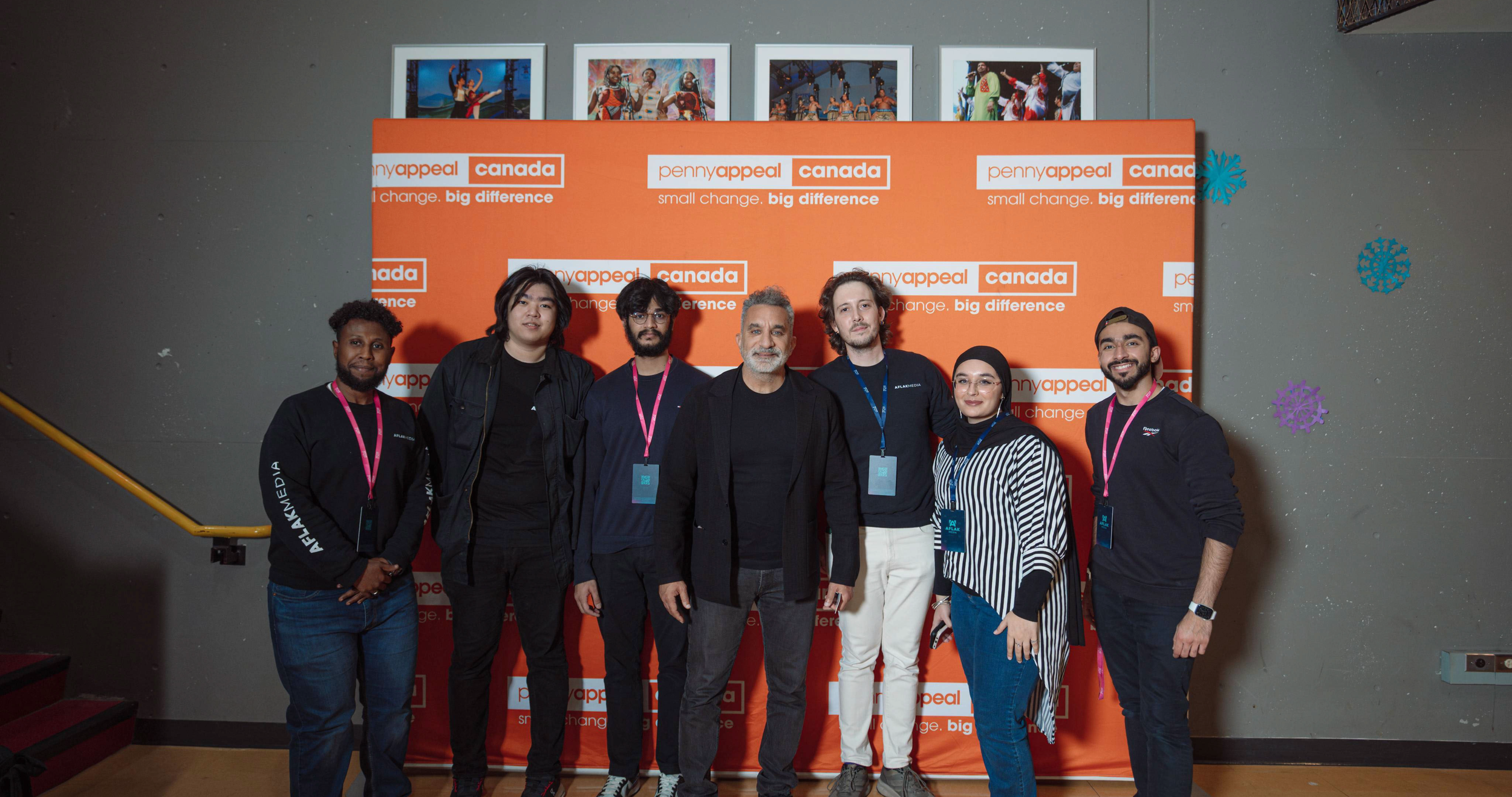 Group of six people standing in front of an orange backdrop with 'pennyappeal canada' logos, some of whom are wearing black 'ALAMDIA' jackets and conference badges.