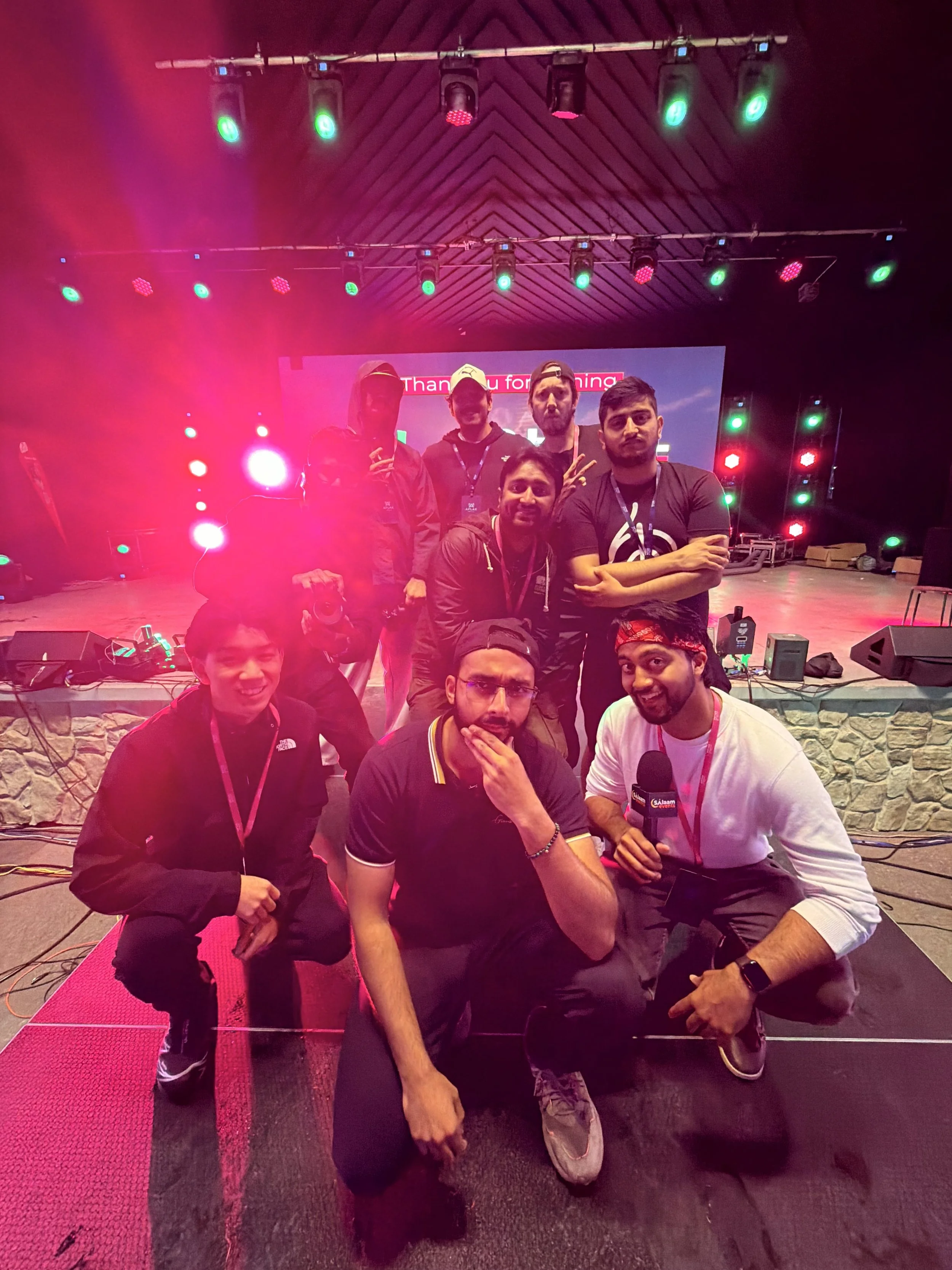 Group of nine young men posing on stage with colorful lights, some smiling and one holding a microphone, in a lively indoor event setting.