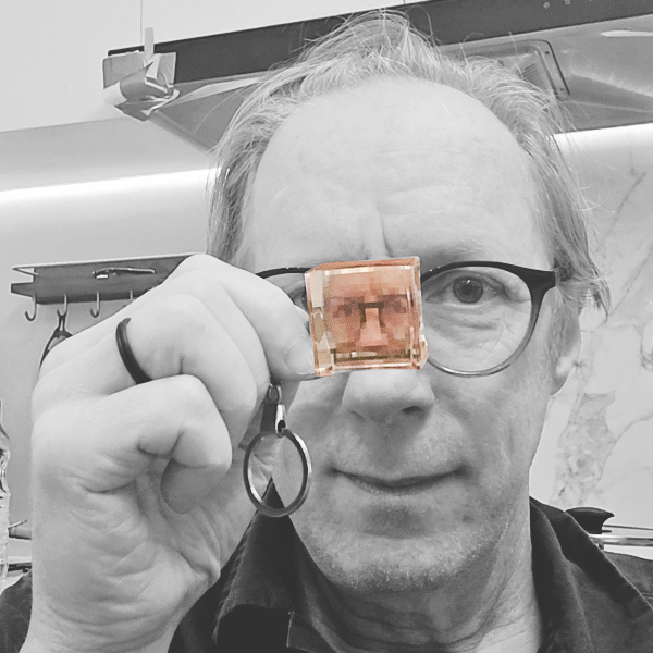 A man with glasses holding a pixelated keychain up to his right eye, with a background that appears to be an office or workshop.