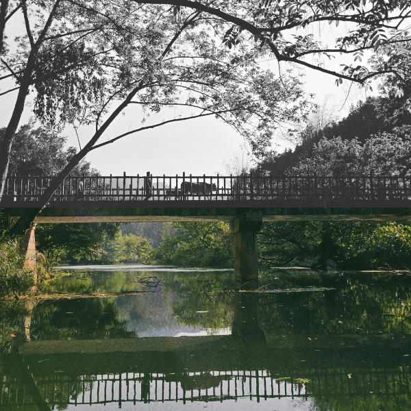 A black and white photo of a bridge crossing over a calm river with trees and a hillside in the background.