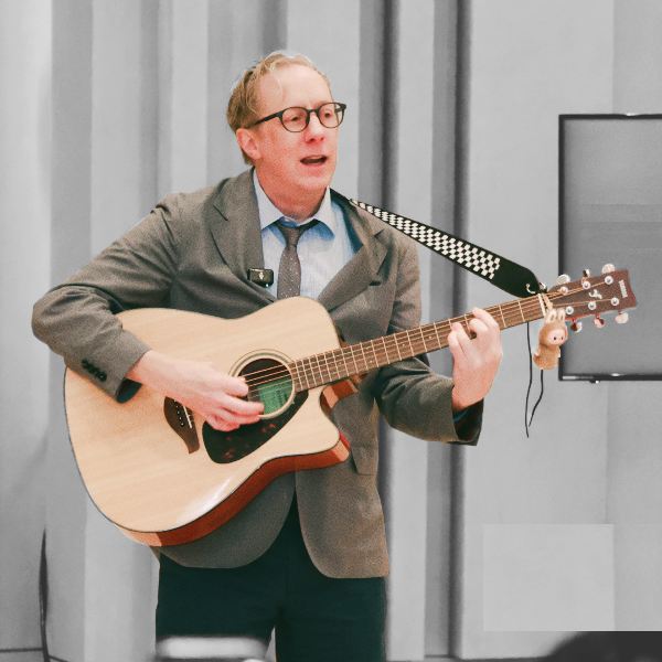 Man in glasses and suit playing acoustic guitar and singing.