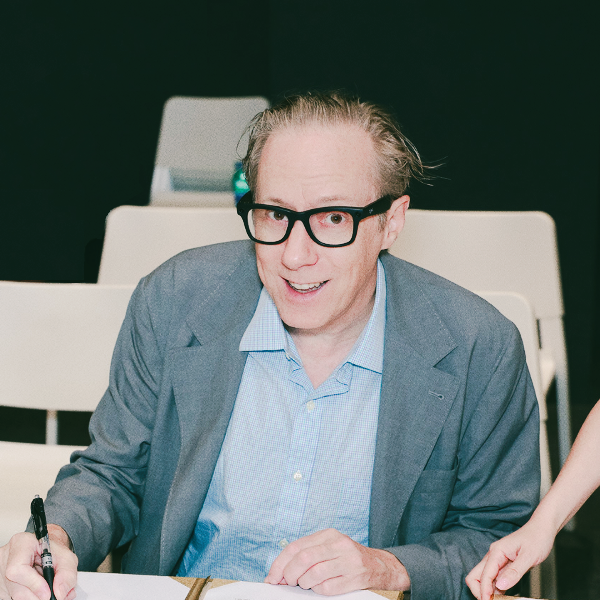 A man with glasses, wearing a gray blazer and a light blue shirt, sitting at a desk with a pen in his hand, smiling at the camera in a meeting or conference room.