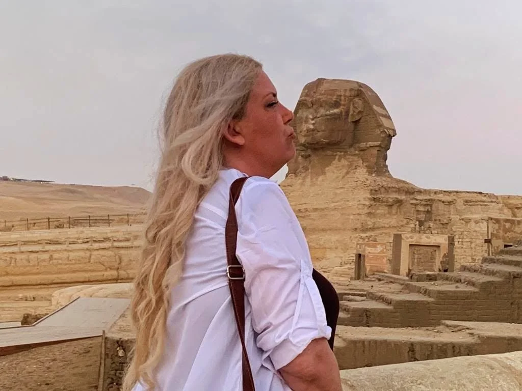 A woman with long blonde hair standing outdoors near the Great Sphinx of Giza, Egypt, during daytime.