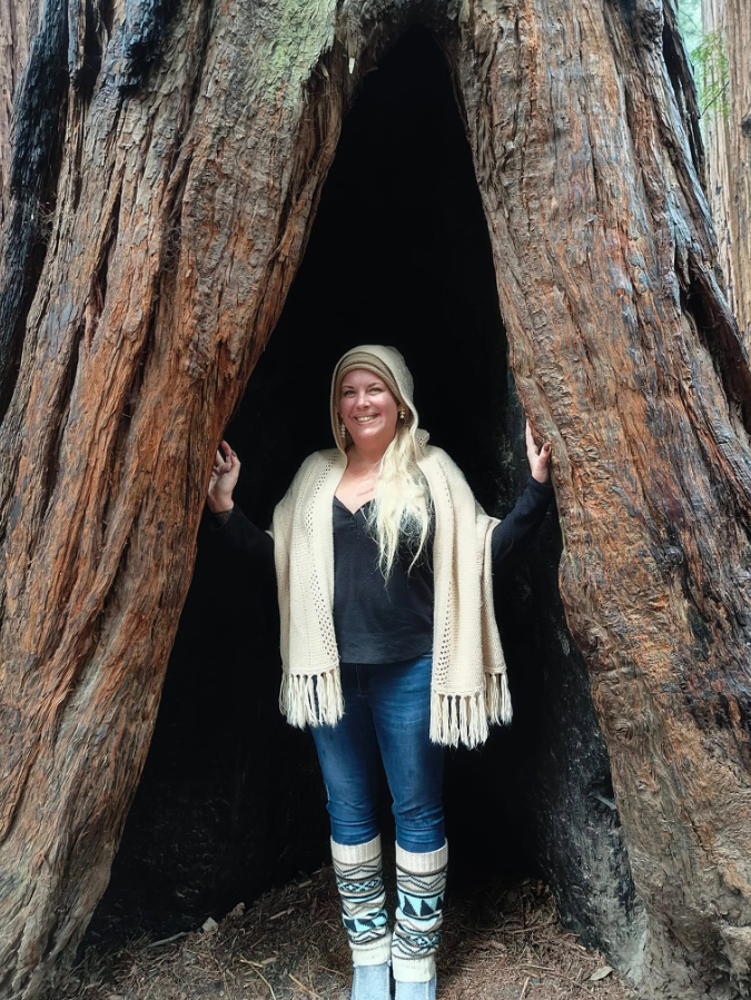 A woman standing inside the hollow of a large redwood tree in a forest, smiling and looking at the camera, wearing a hat, scarf, and patterned socks.