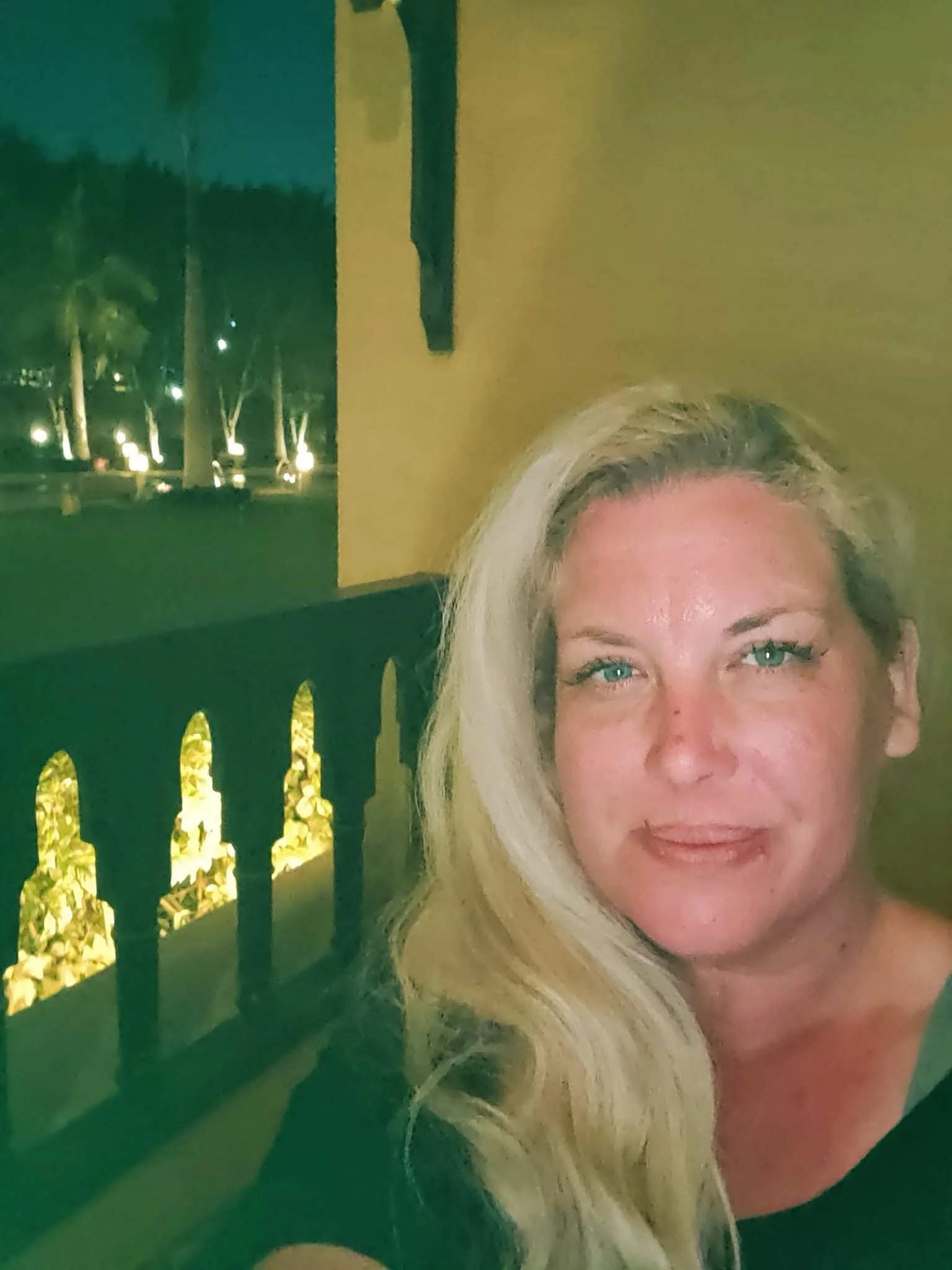 Close-up selfie of a woman with long blonde hair and blue eyes, partially visible, taken outdoors at night near a yellow wall with a balcony railing, illuminated by warm yellow lights.