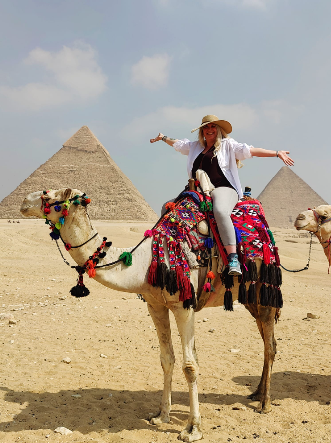 Woman with arms outstretched riding a camel decorated with colorful textiles and tassels in front of pyramids in Egypt.
