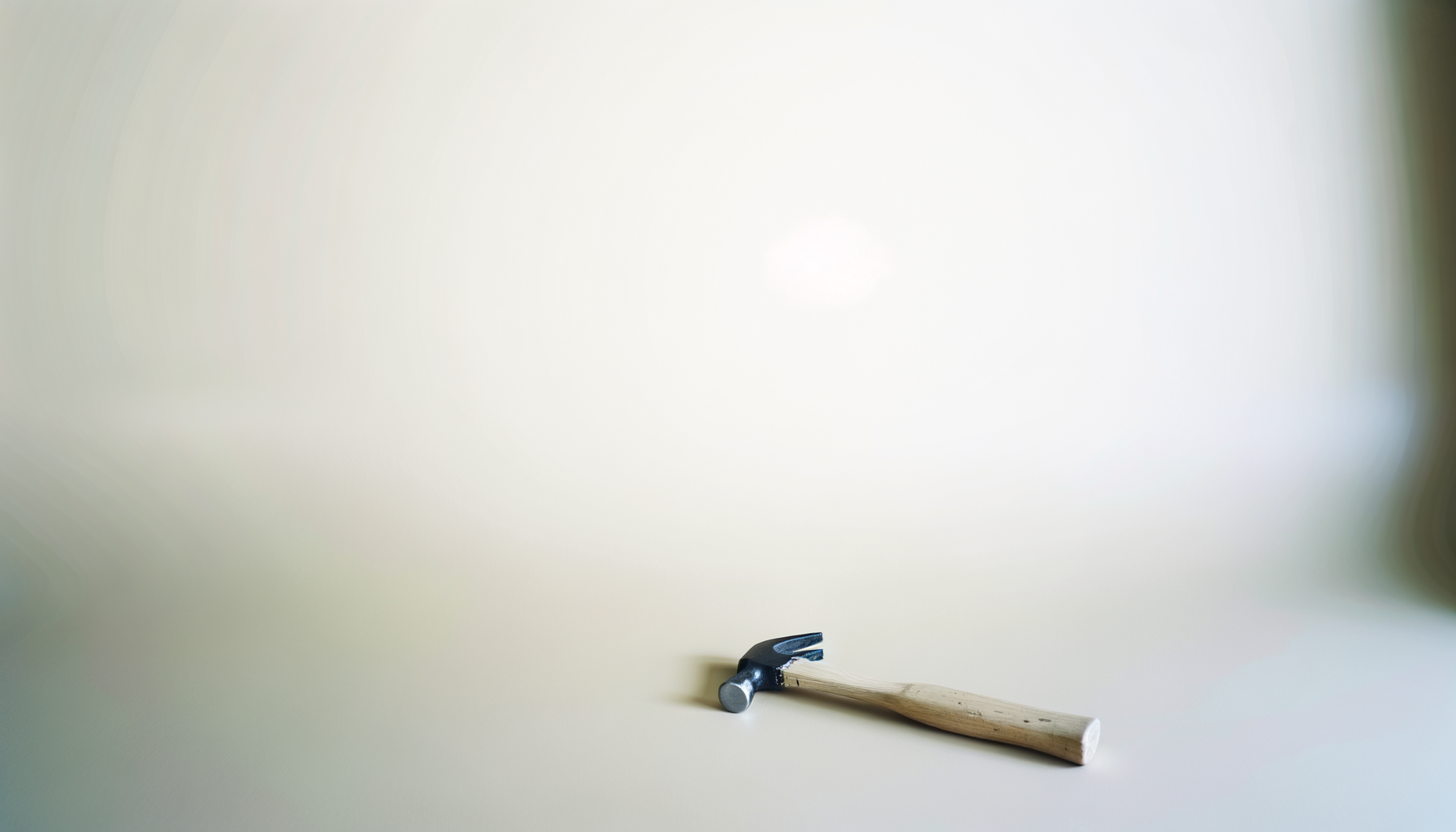 A hammer with a black head and a wooden handle resting on a light-colored surface against a plain, slightly gradient background.