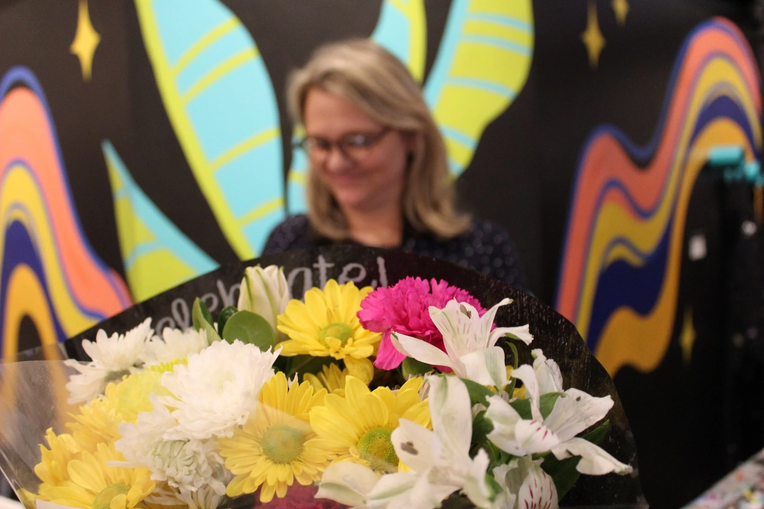 A colorful bouquet of yellow, white, and pink flowers held in front of a person with blonde hair, glasses, and a black polka dot shirt, standing in front of a vibrant, abstract mural with bold patterns and stars.