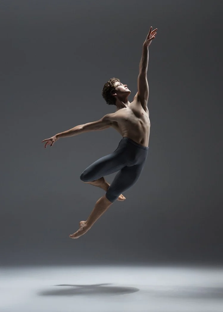 A male ballet dancer jumps mid-air with one arm extended upward and the other extended to the side, in a dark studio setting.