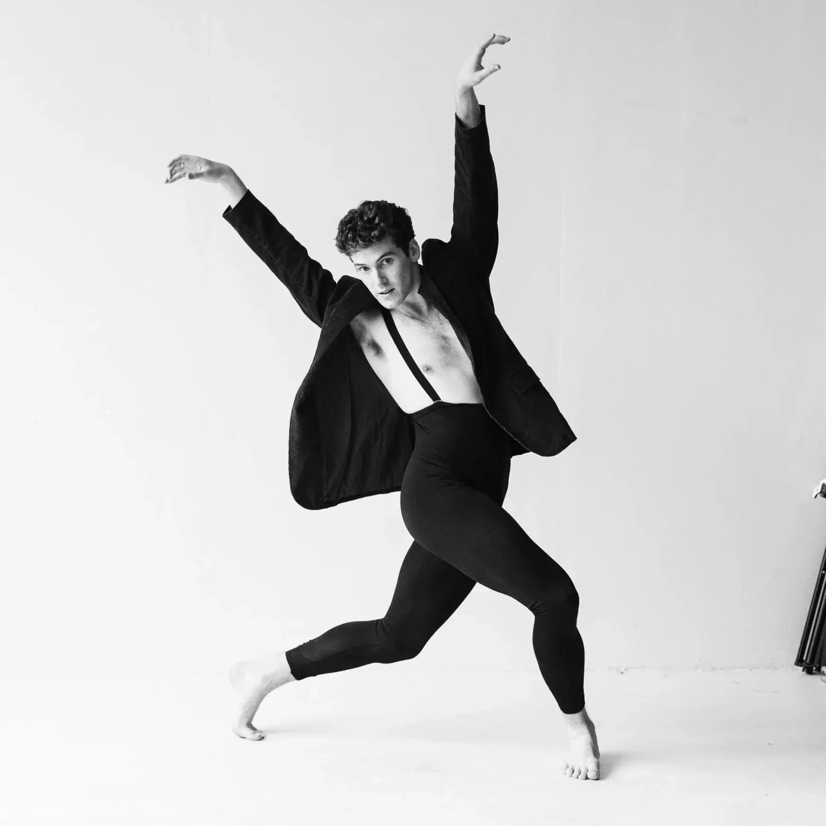 A person performing a dance pose in a studio with a plain background, wearing black clothing and a blazer with partially open shirt. The person has curly hair and is balancing on one leg with arms raised.