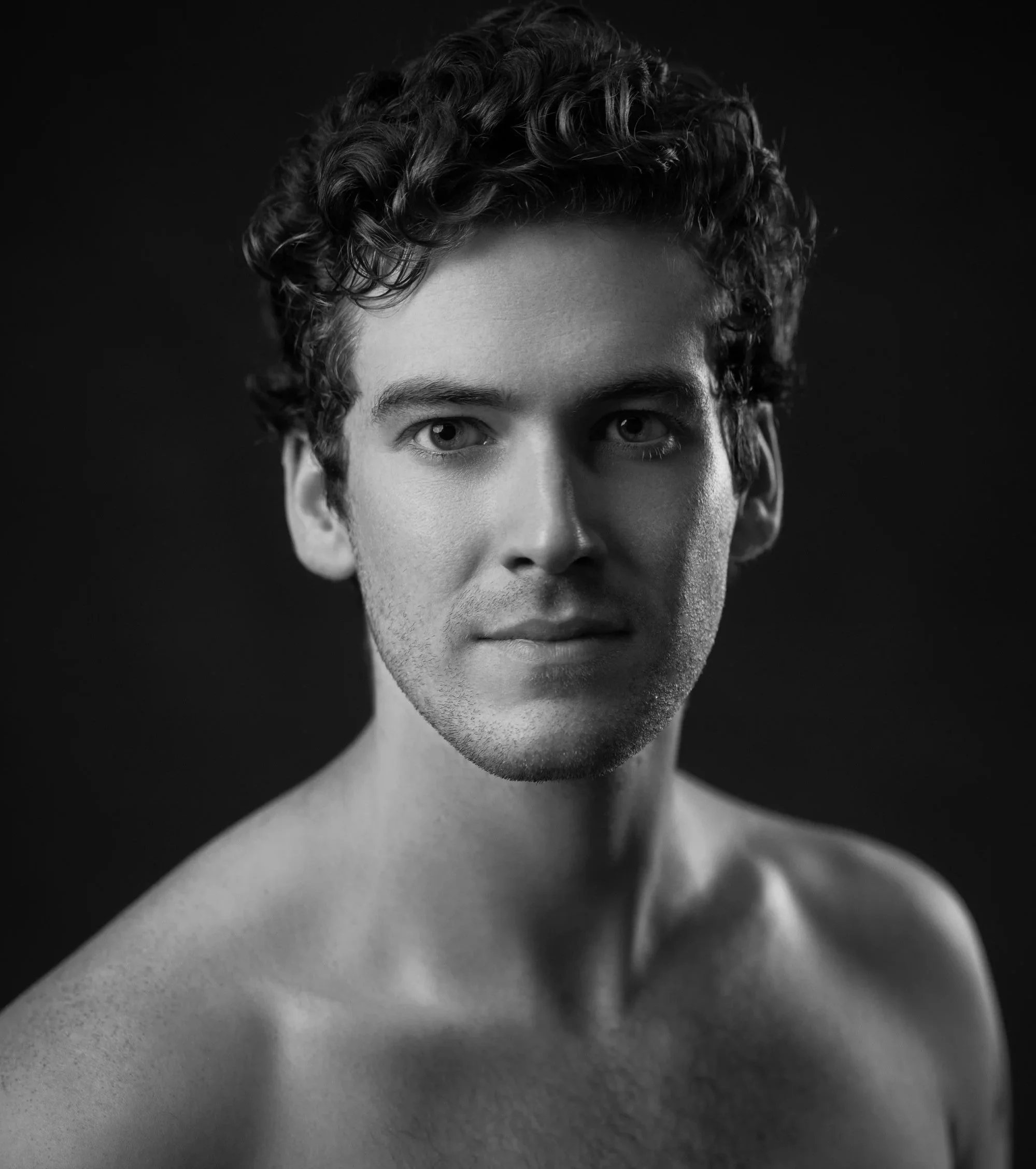 Close-up black and white photograph of a young man's face and upper body, showing curly hair and intense eyes, with a dark background.