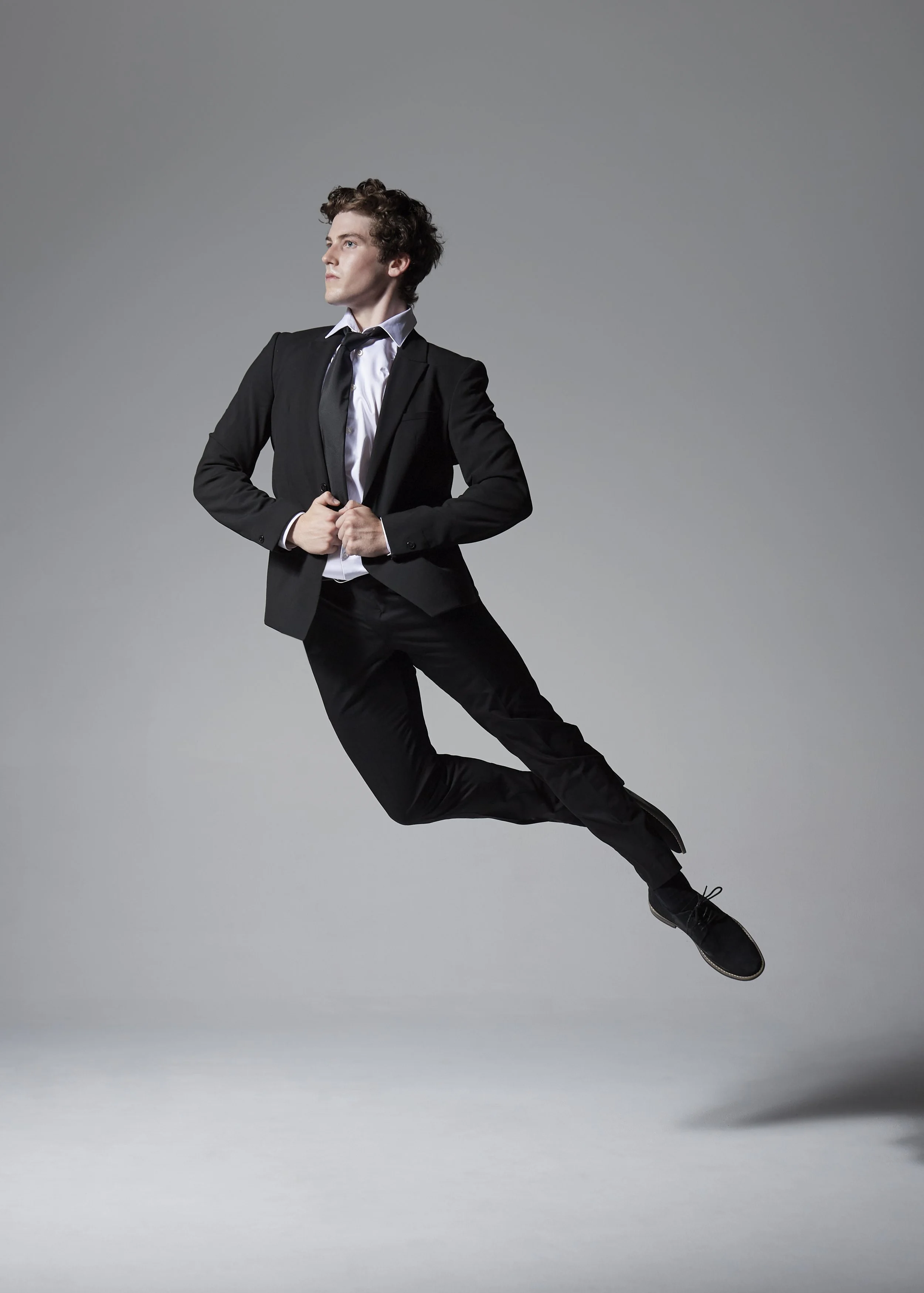 A man in a black suit, white shirt, and black tie is floating mid-air against a gray background, holding the lapels of his jacket with both hands.