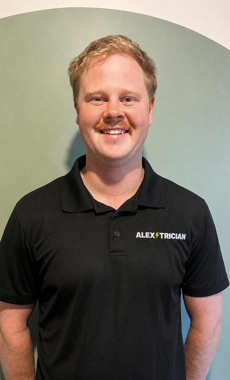 A young man with red hair, a mustache, and light skin smiling, wearing a black polo shirt with the name 'ALEX TRICIAN' embroidered on it, standing in front of a two-tone background.