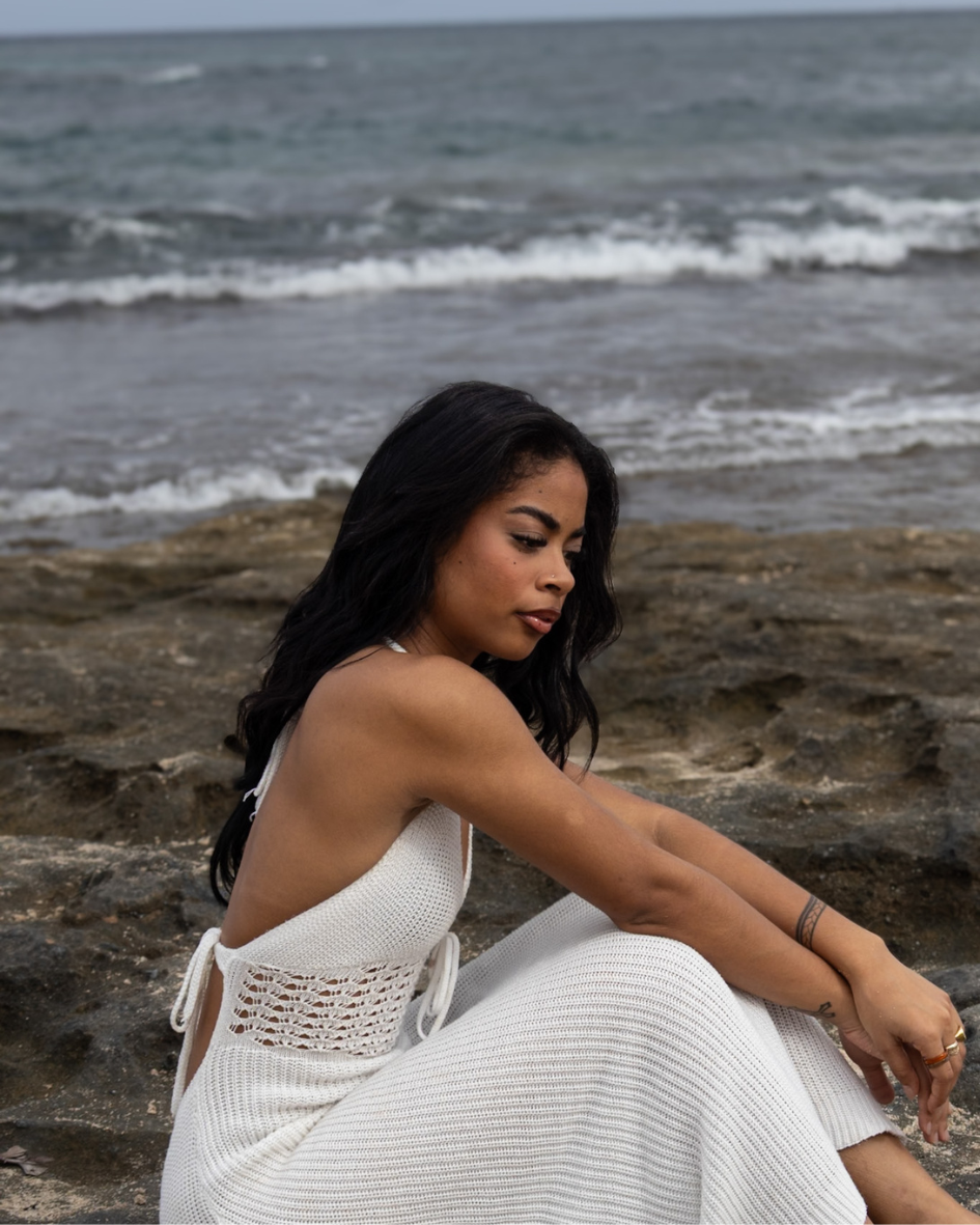 A woman with dark hair sitting on rocks at the beach, wearing a white sleeveless dress, with ocean waves in the background.