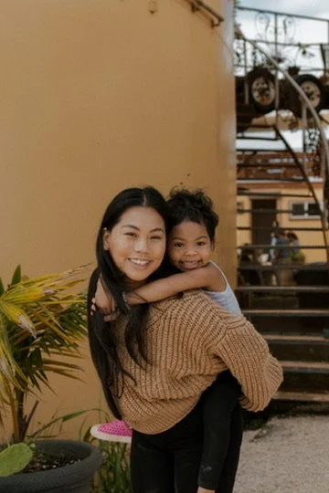 A woman holding a young girl outside near a staircase with a yellow wall and a potted plant nearby.