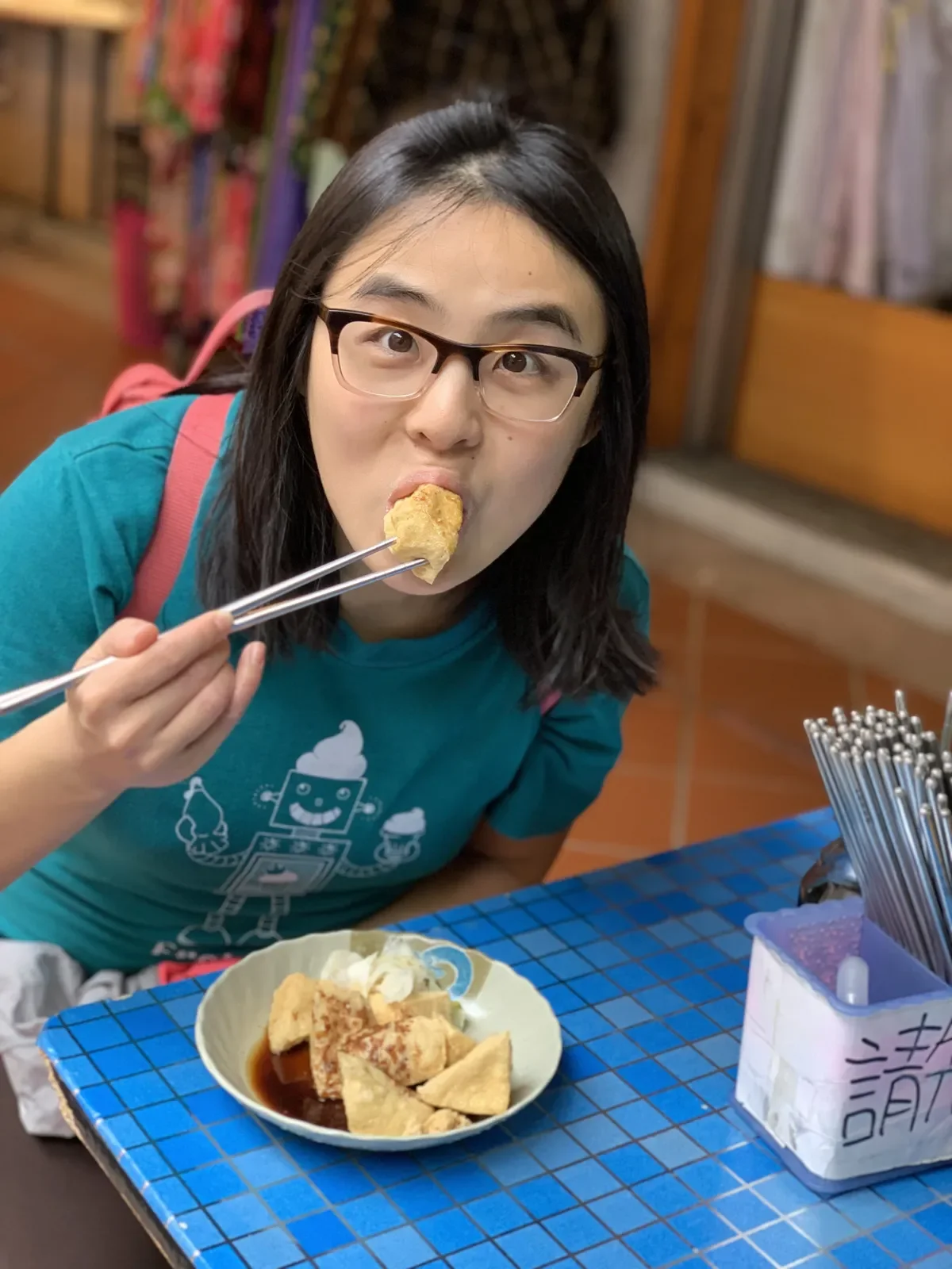 Shirley wearing a teal shirt, pink backpack, eating stinky tofu with metal chopsticks