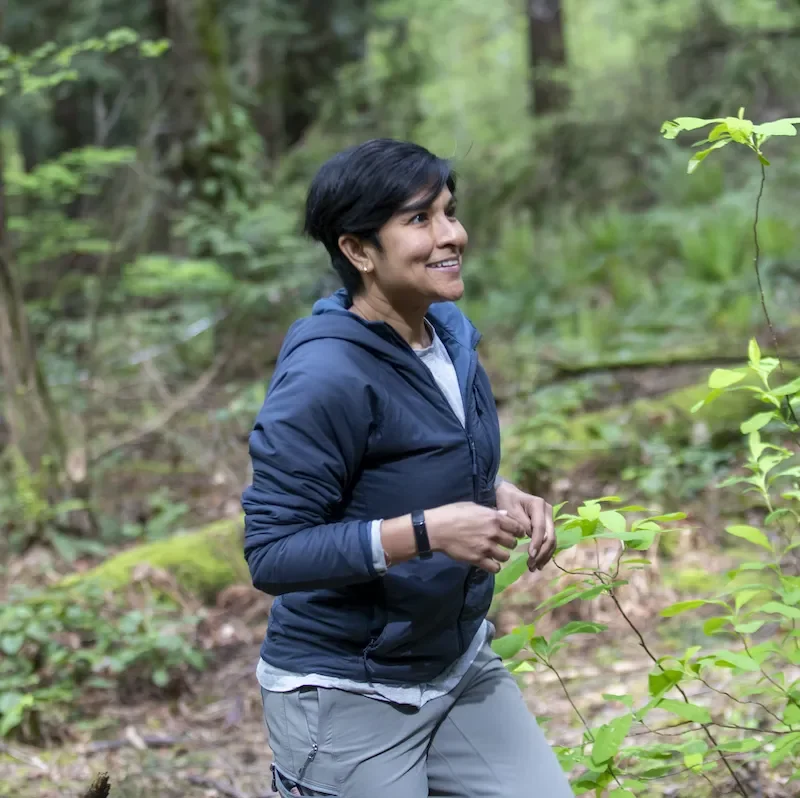 Meera under a forested canopy, with young plant shoots surrounding her and a mossy log behind