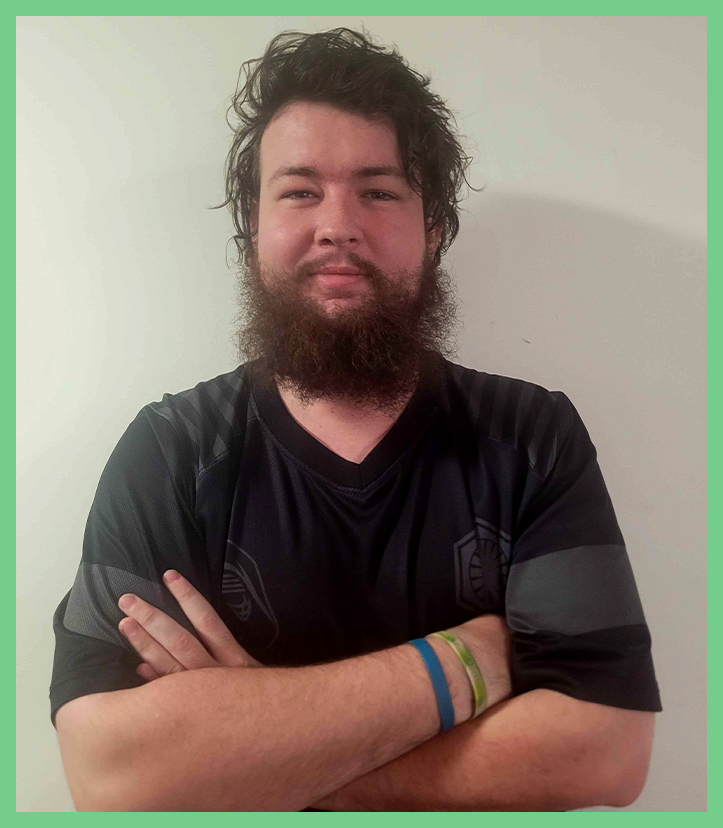 A young man with dark, messy hair and a full beard, standing with arms crossed, wearing a black sports jersey, against a plain white wall.