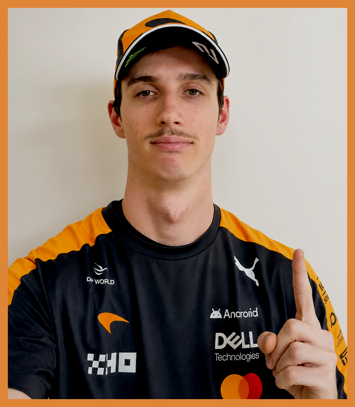A man in a racing team uniform with sponsor logos, wearing a yellow and black cap, making a 'number one' gesture with his right hand, standing against a plain white background.
