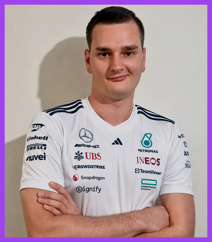 A young man with short dark hair, wearing a white Mercedes-AMG Petronas Formula One Team shirt with multiple sponsor logos, standing with his arms crossed against a plain wall.