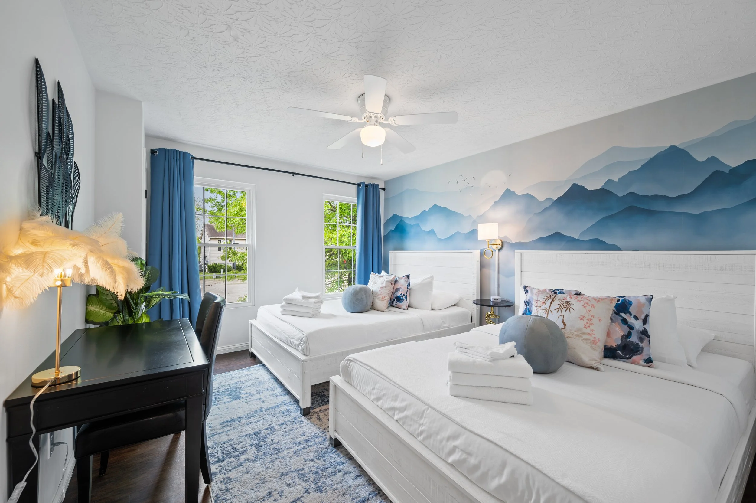 A bright bedroom with a mountain mural on the wall, two white beds with decorative pillows, a ceiling fan, windows with blue curtains, a black desk with a lamp, and a blue patterned rug.
