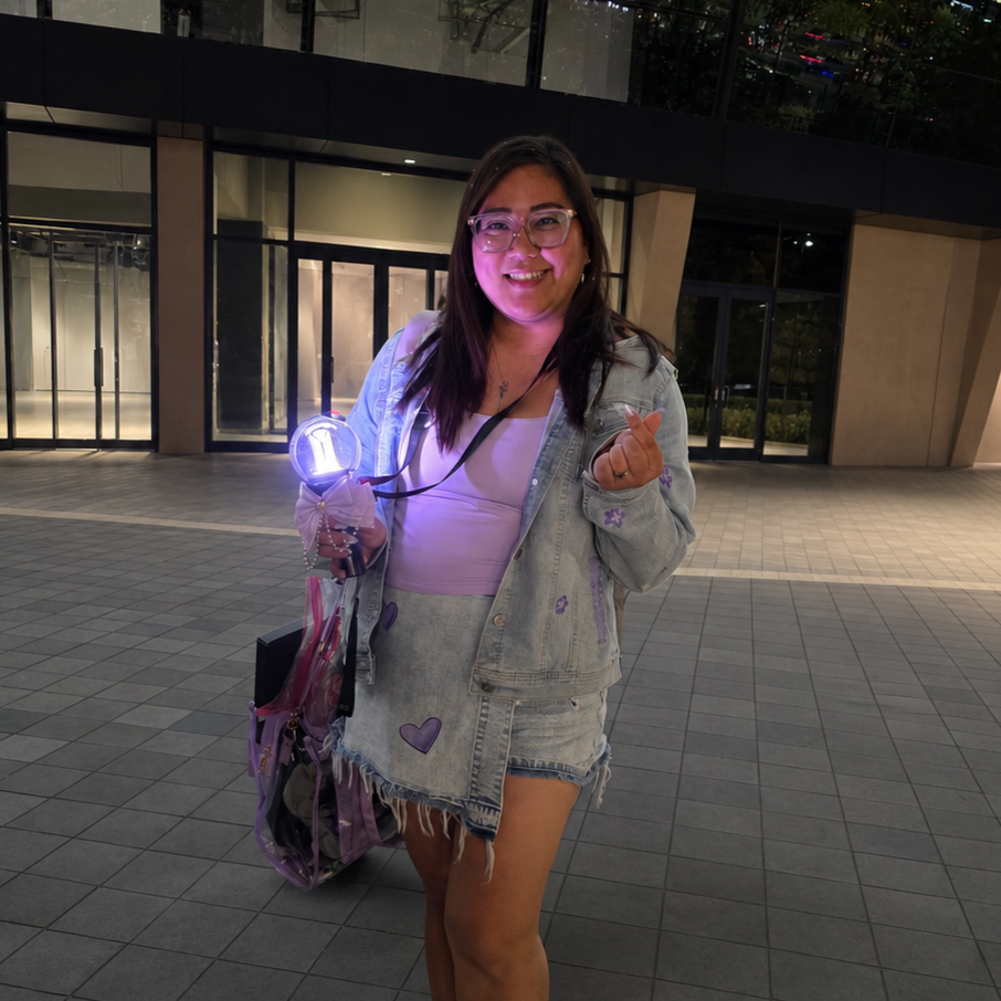 A smiling woman with glasses in a denim jacket and skirt standing outside at night, holding a purple light and making a finger heart gesture.
