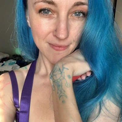 Young woman with vibrant blue hair, light skin, and blue eyes, smiling at the camera, wearing a purple tank top, indoors.