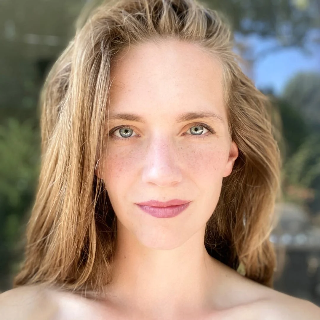 Close-up portrait of a young woman with long, wavy red hair and blue eyes, outdoors with blurred greenery and sky in the background.