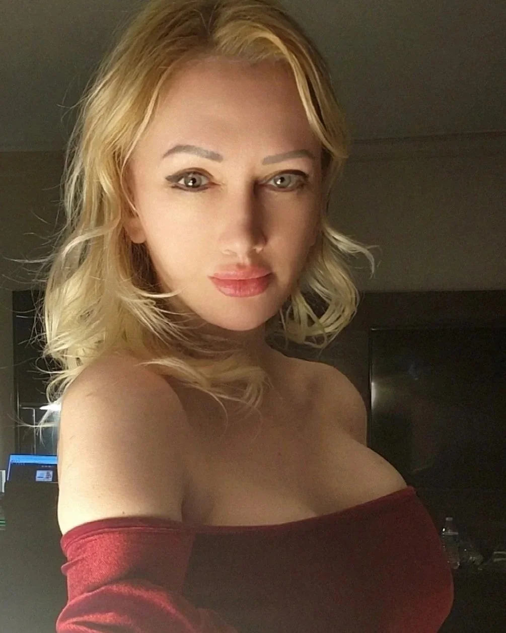 A woman with blonde, shoulder-length hair and light skin taking a selfie in a dimly lit room, wearing an off-the-shoulder red top.