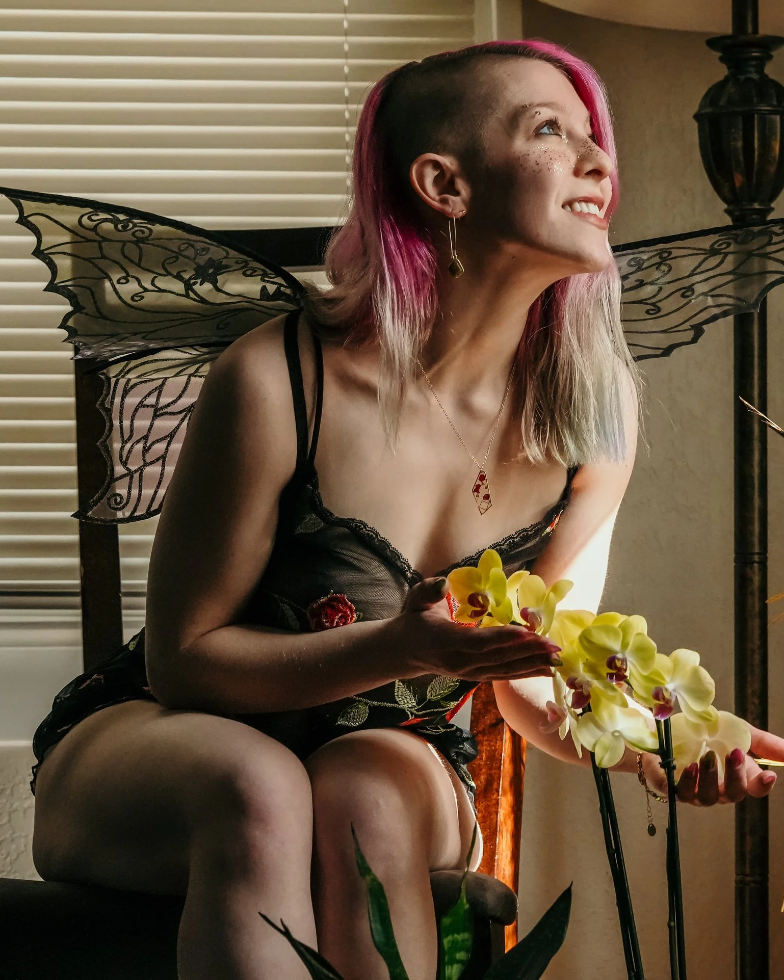 A woman with pink and blonde hair, fairy wings, and wearing black lace clothing smiles while holding yellow orchids in a dimly lit room.