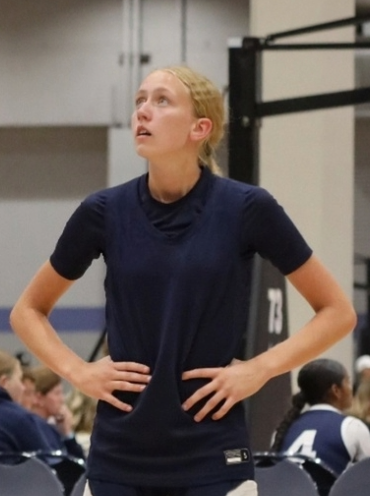 A young woman with blonde hair, wearing a navy sports uniform, stands with her hands on her hips and looks upward seconds after a volleyball game.