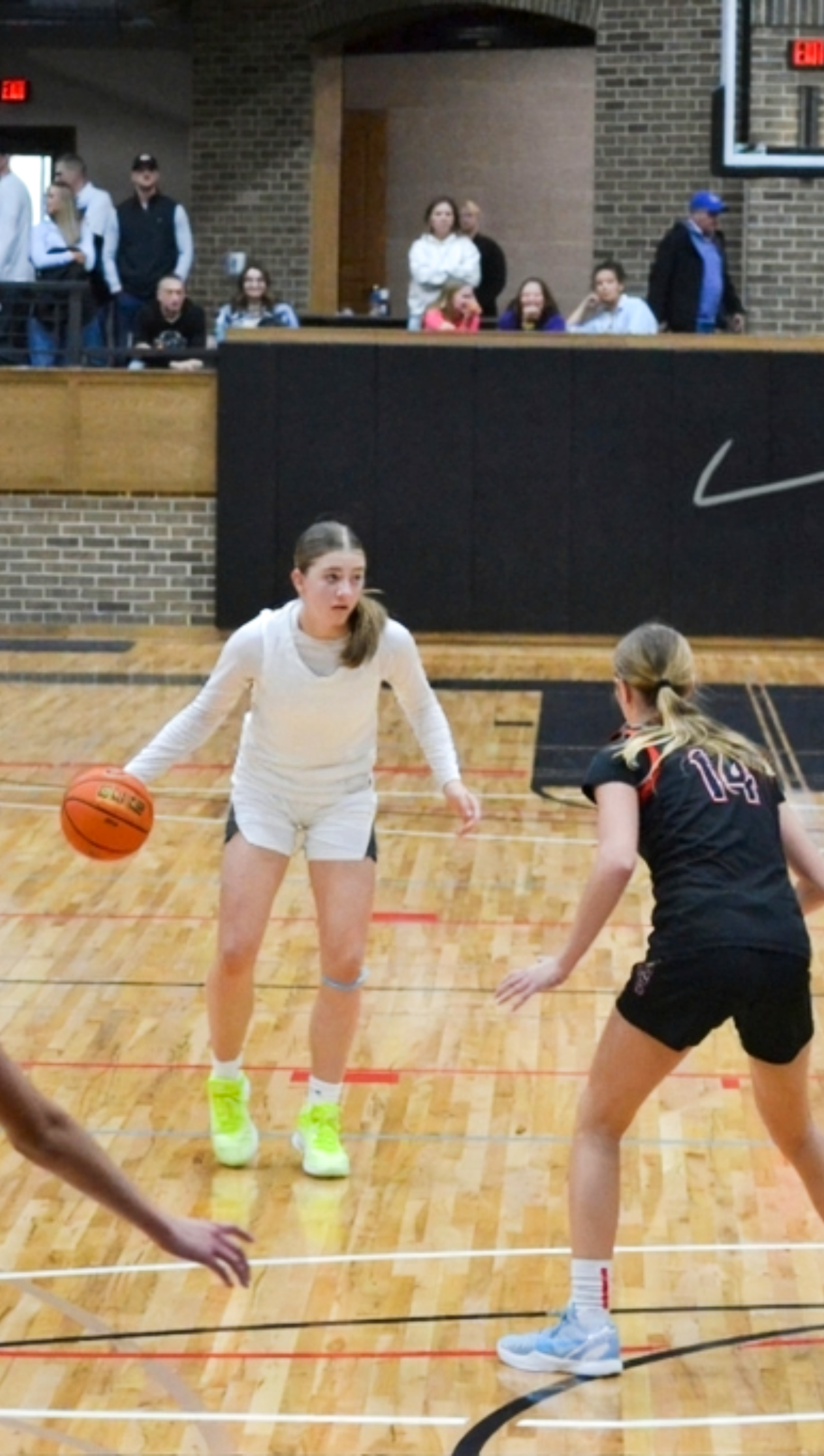Two female basketball players on the court during a game, one in a white uniform dribbling the ball and the other in a black uniform defending, with people watching from the bleachers in the background.