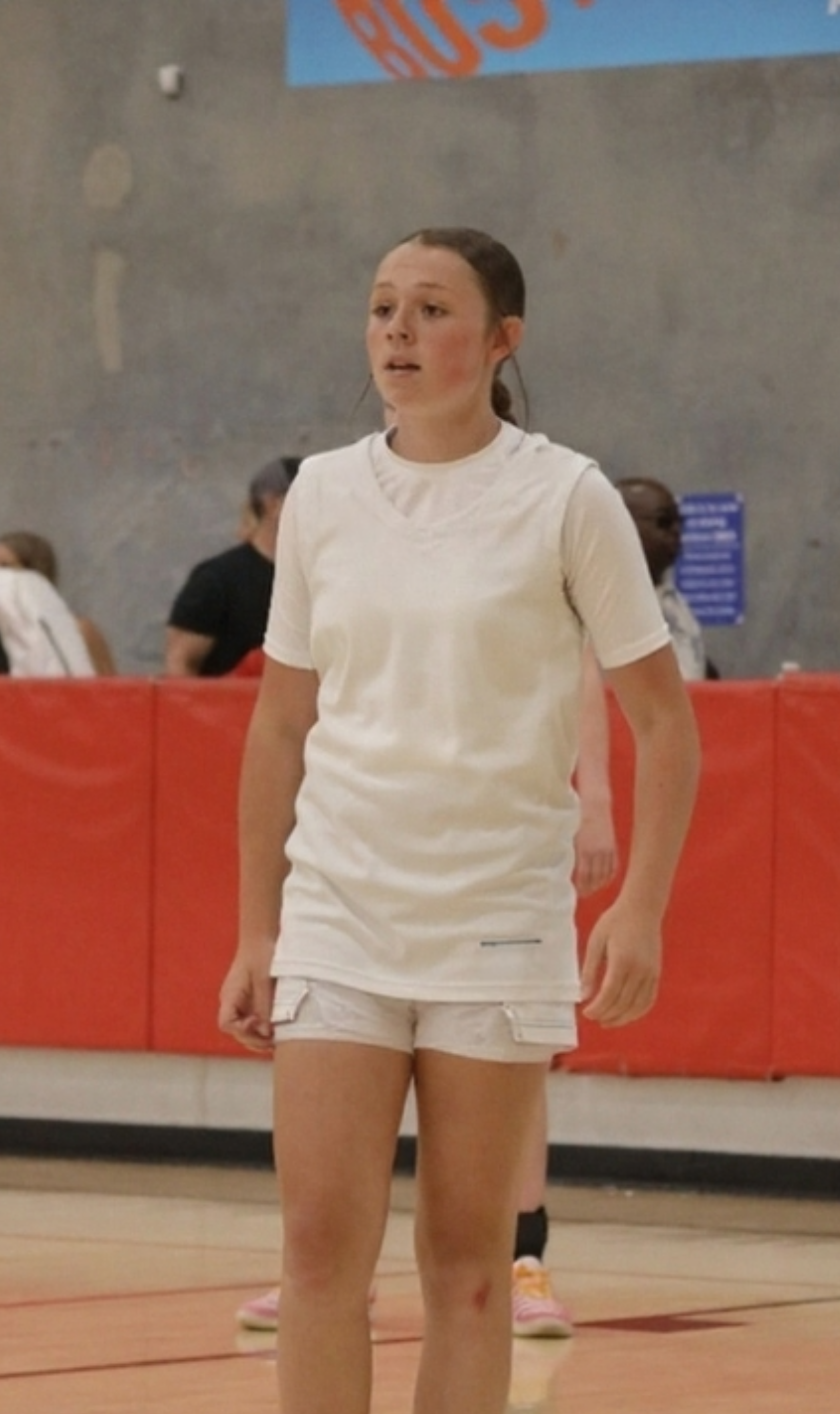 A young girl in a white sports outfit, standing on a gymnasium floor, with other children and a red padded barrier in the background.