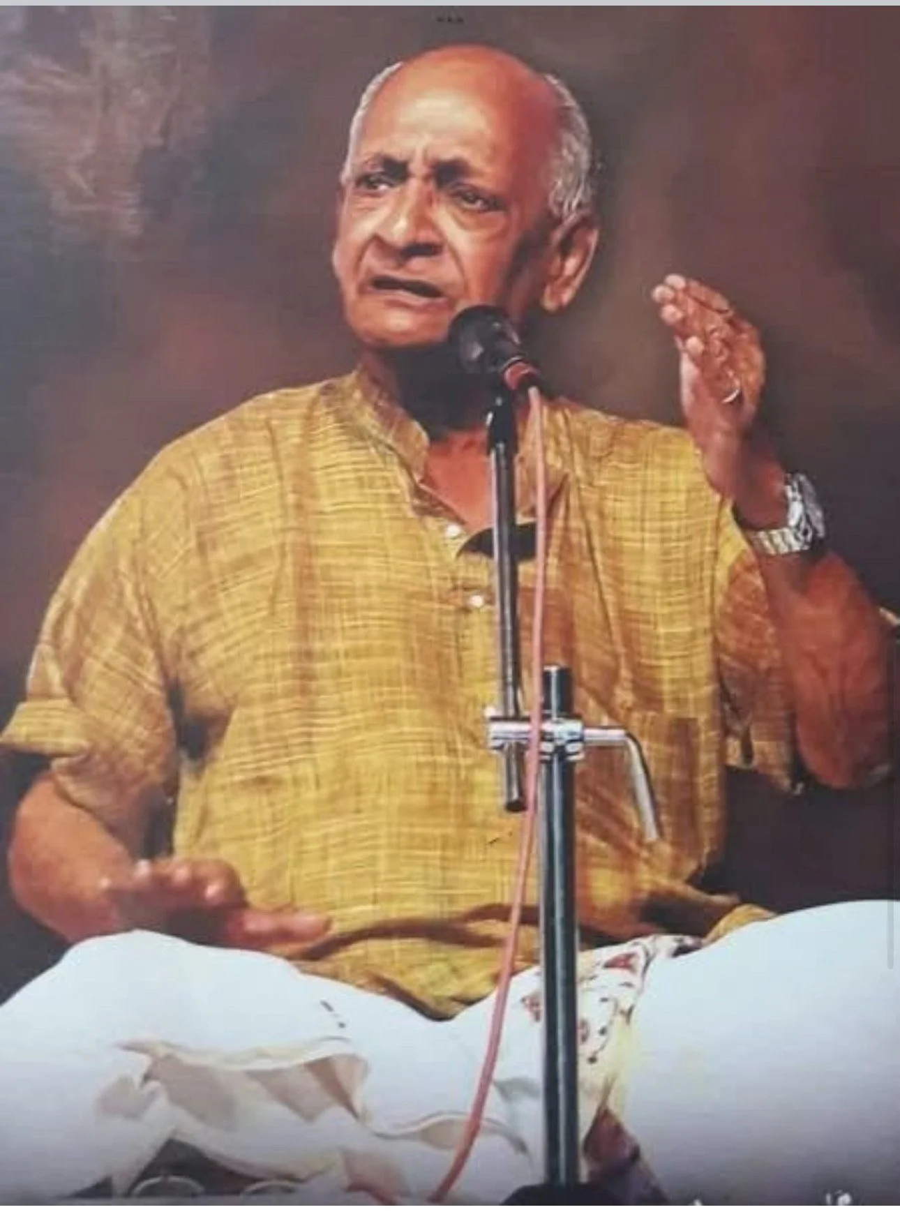 An elderly man with a shaved head, wearing a yellow checkered shirt, is speaking into a microphone while making a hand gesture. He appears to be giving a speech or address, and is seated on a white cloth with some fabric draped in front of him.