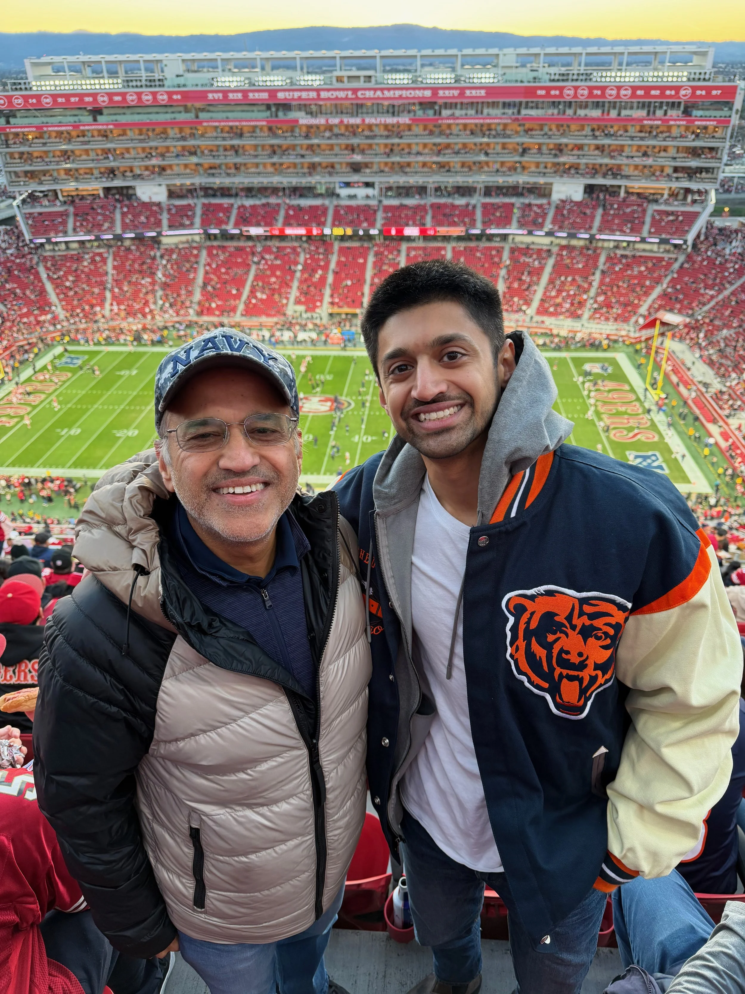 Two smiling men standing in a stadium with a football field in the background, celebrating a game, with fans in the stands, during sunset.