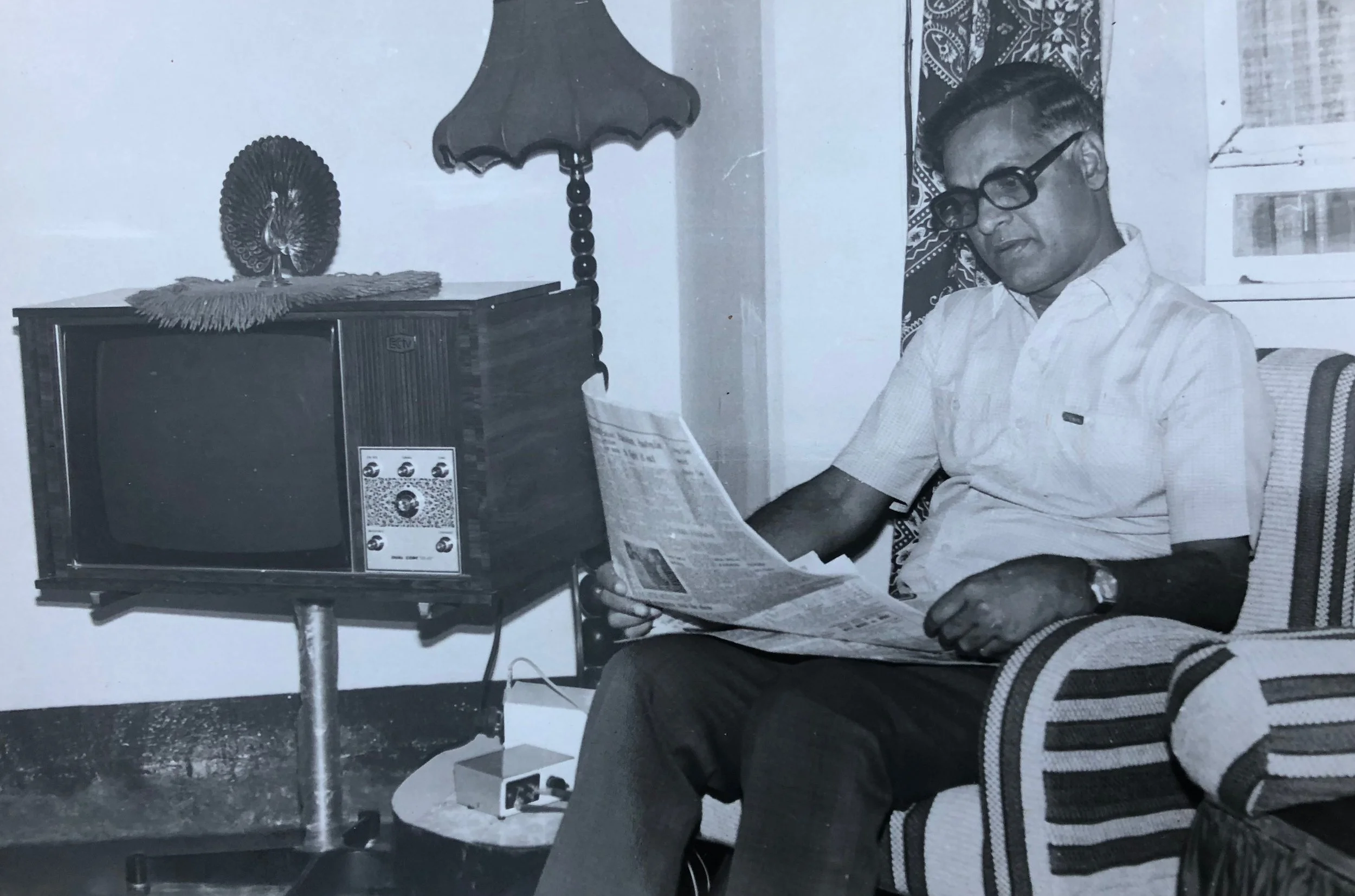A man with glasses sitting in a chair reading a newspaper, with a vintage television set on a small table next to him, in a room with patterned curtains.