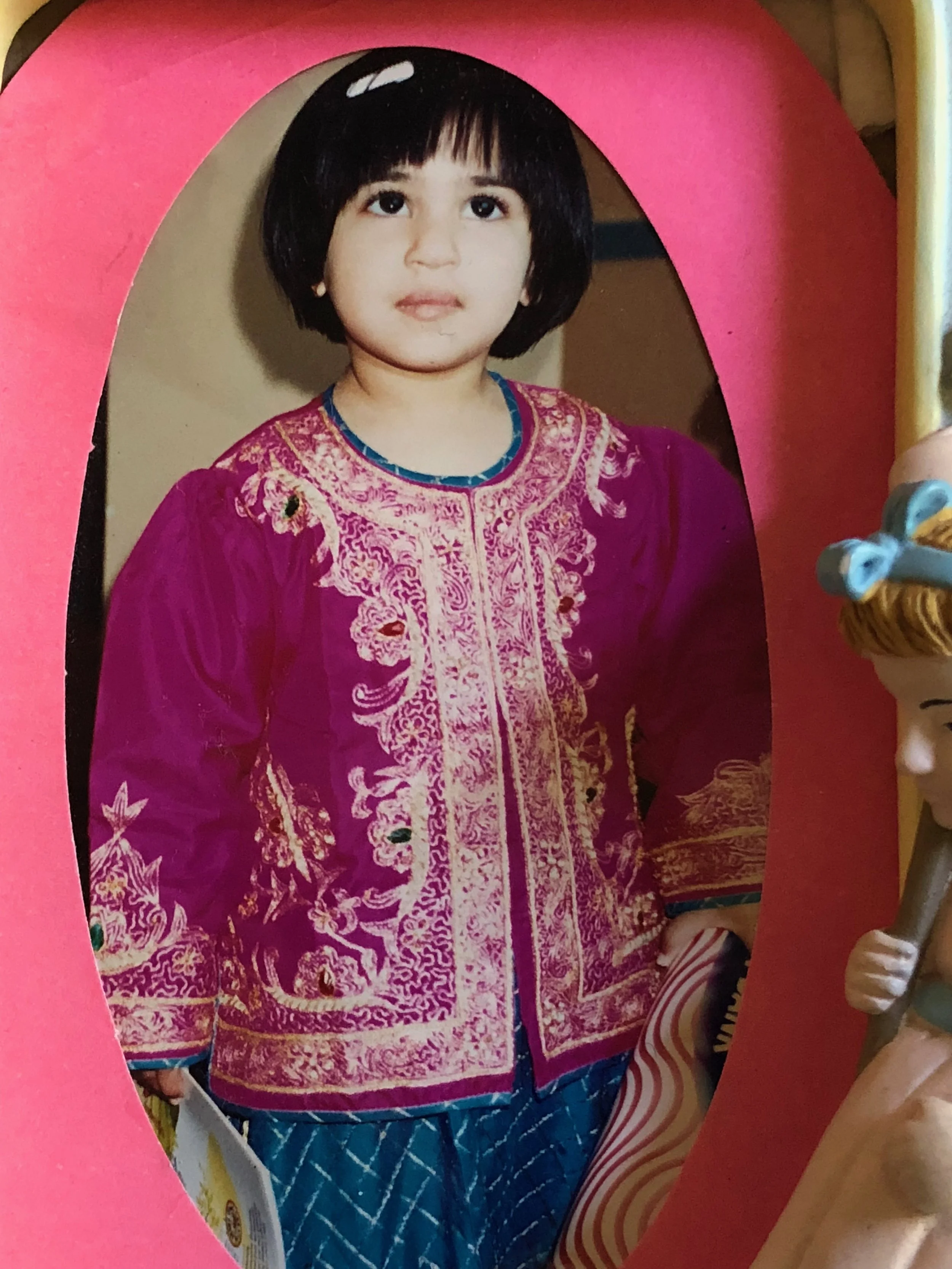 A girl with short black hair and big eyes, wearing a traditional pink and gold embroidered dress, standing inside a pink and yellow cardboard photo frame.