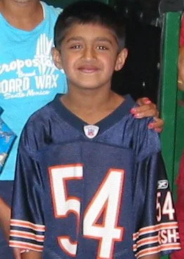A young boy smiling, wearing a navy blue football jersey with the number 54, standing next to a person in a blue t-shirt.