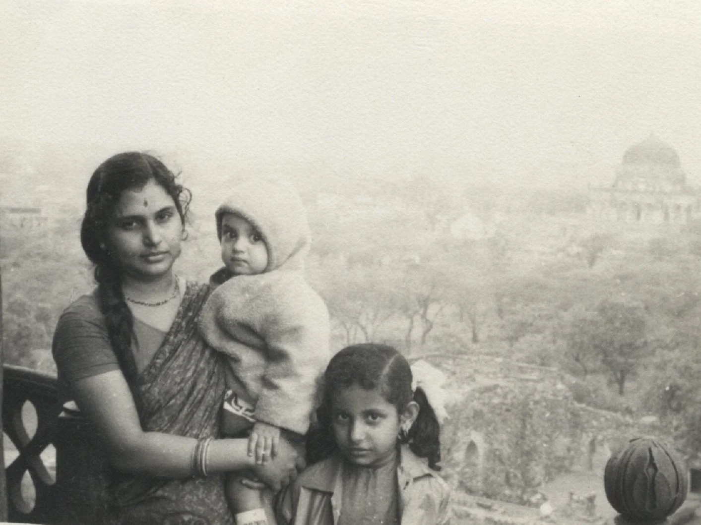 A black and white photo of a woman holding a young child, with another girl standing beside them. The background features a distant landscape with trees and a building.