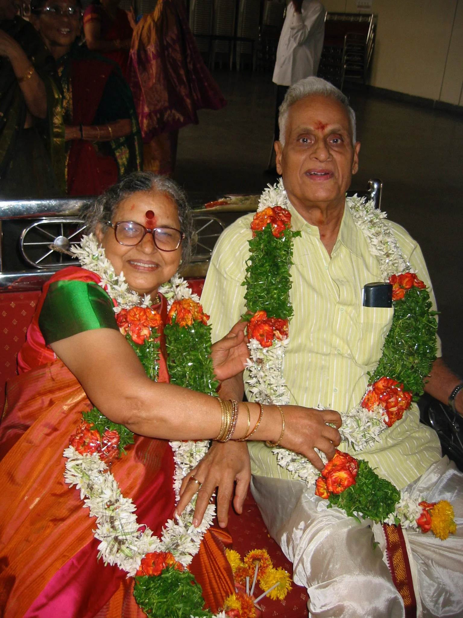 An elderly couple dressed in traditional Indian attire, wearing garlands and jewelry, celebrating at a cultural event, sitting closely together, smiling, and holding hands.