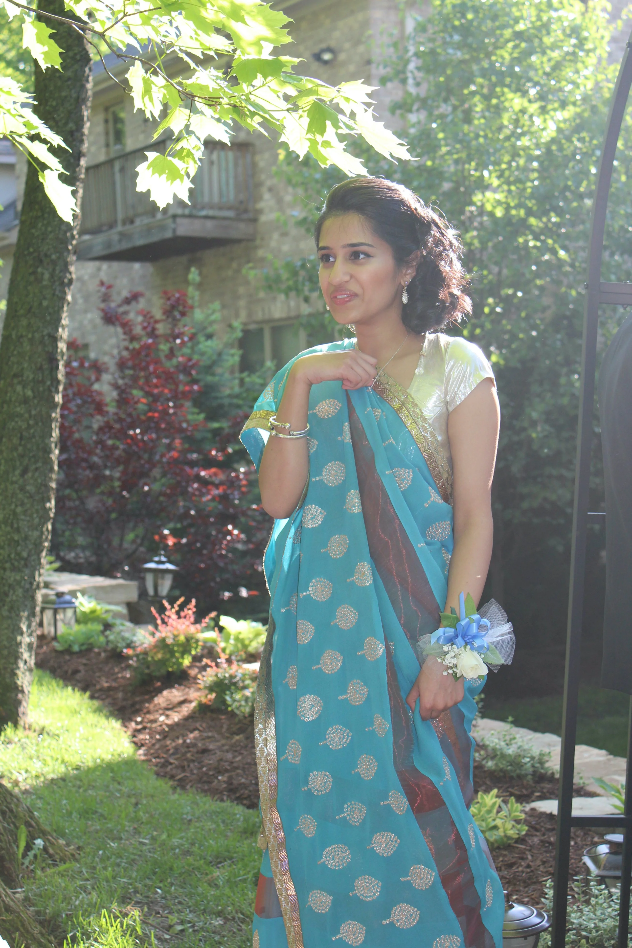 A woman in a traditional blue saree with gold accents standing outdoors in a garden, holding a small bouquet with white flowers and a blue ribbon, with trees and a stone building in the background.