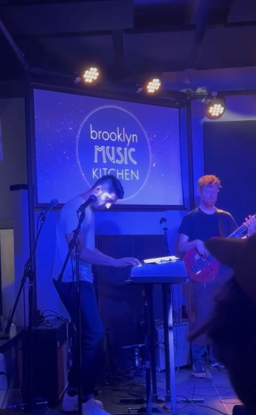 Musicians performing on stage at Brooklyn Music Kitchen with a blue-lit backdrop and logo projection.