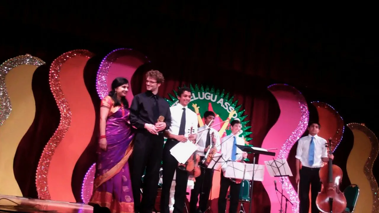 Group of people on stage at an event with musical instruments and colorful backdrop reading 'ELUGU AASAP'